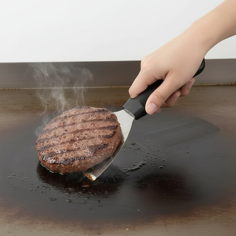 KRD Pro Stainless Steel Griddle Scraper Spatula 25cm lifting a grilled burger on a flat griddle surface.