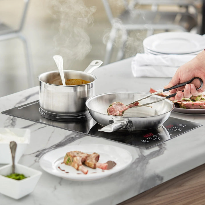 Buffet induction warmer with cookware on marble surface, featuring steaming pots and plating of cooked dishes.